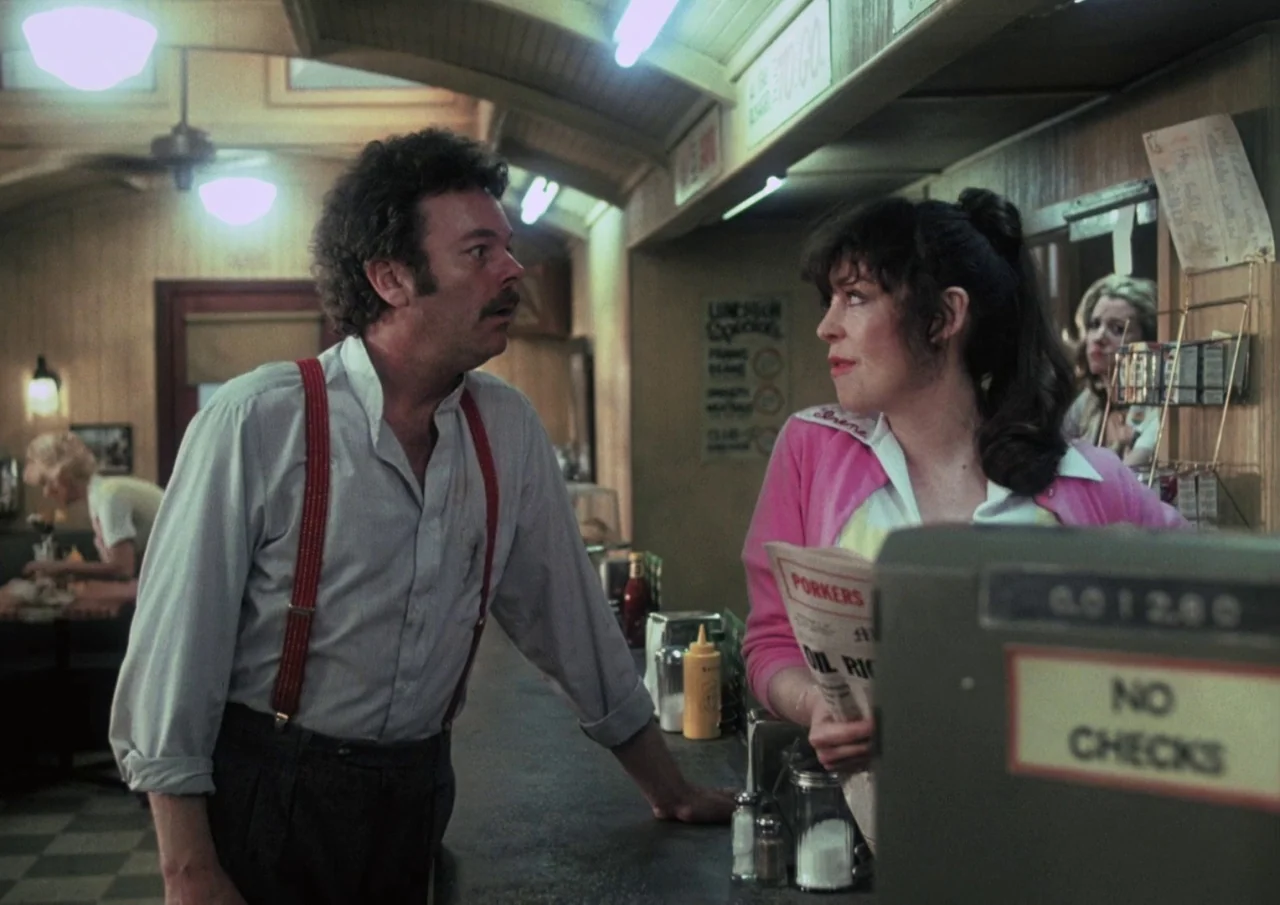 Human Highway (1982) movie still: medium — A 1980s diner interior features wood-paneled walls and a tiled floor. A man with red suspenders speaks to…