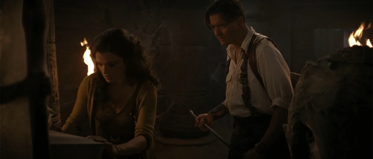 The Mummy Returns (2001) movie still: medium — A dimly lit ancient stone chamber features carved walls and flickering wall torches. A woman studies…
