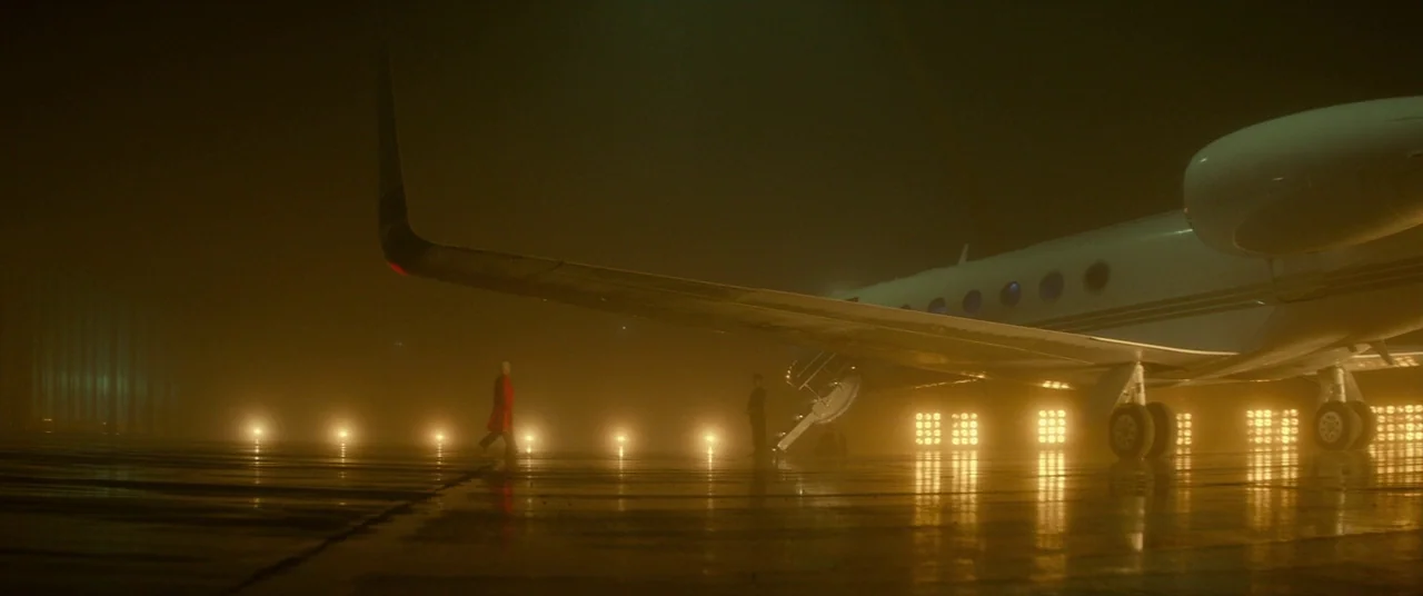 Atomic Blonde (2017) movie still: wide — An airplane sits on a dark, wet tarmac illuminated by a line of floodlights. A lone person wearing a red…