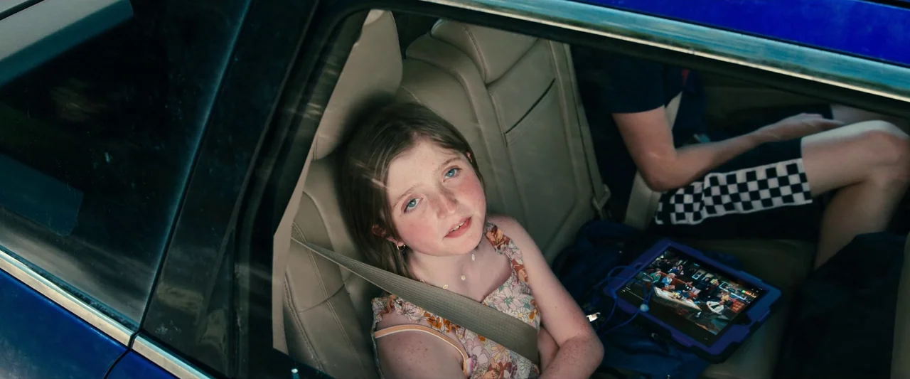 Leave The World Behind (2023) movie still: medium — A view from the exterior of a moving car through the back passenger window. A young girl sits…