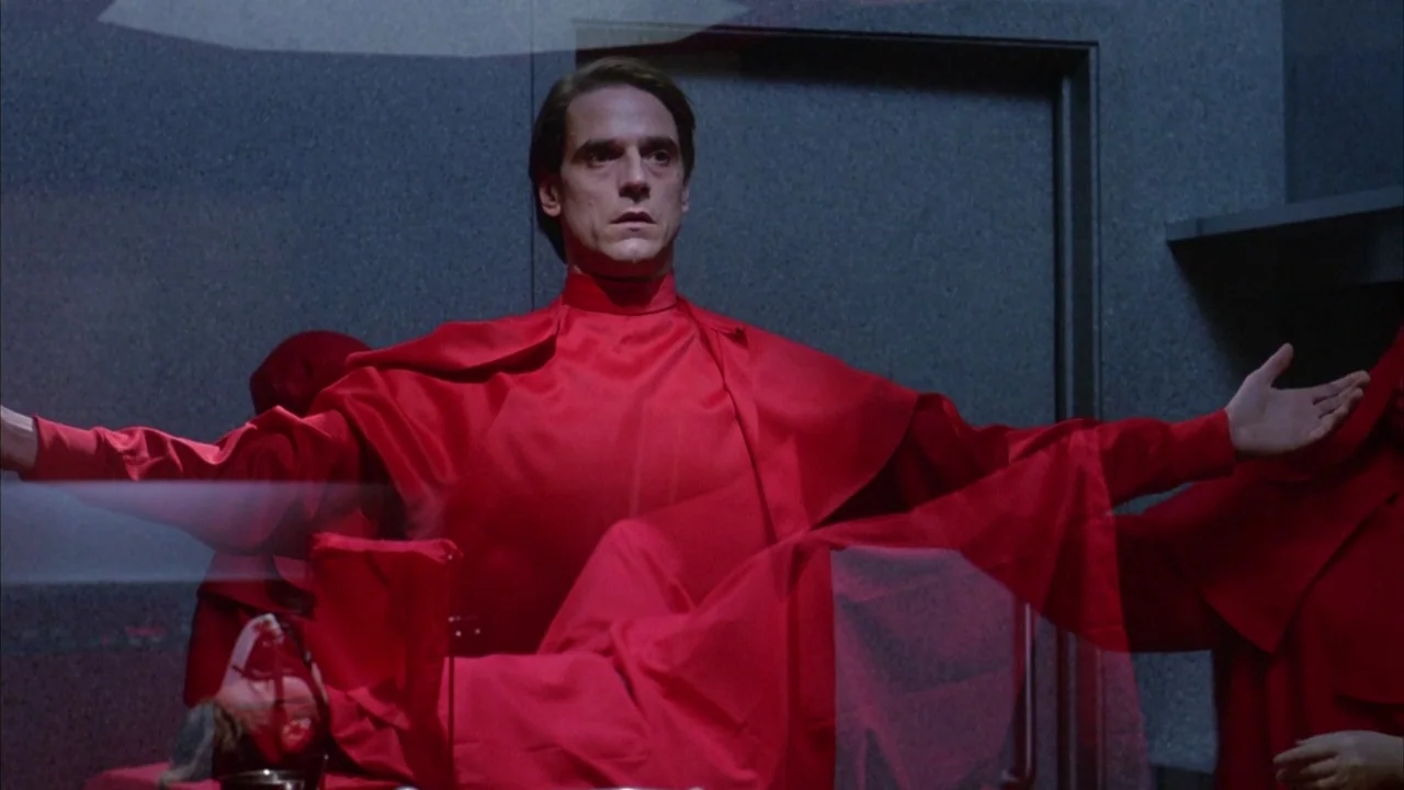 Dead Ringers (1988) movie still: medium — A clinical surgical suite features grey walls and a dark, sterile metallic environment. A man in a red…