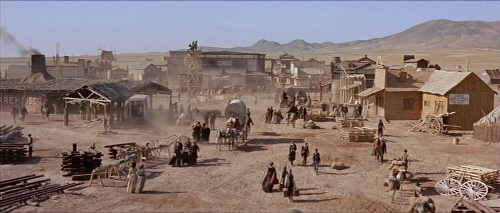 Once Upon A Time In The West (1968) movie still: establishing — A bustling 19th-century frontier town street features wooden structures, rail…