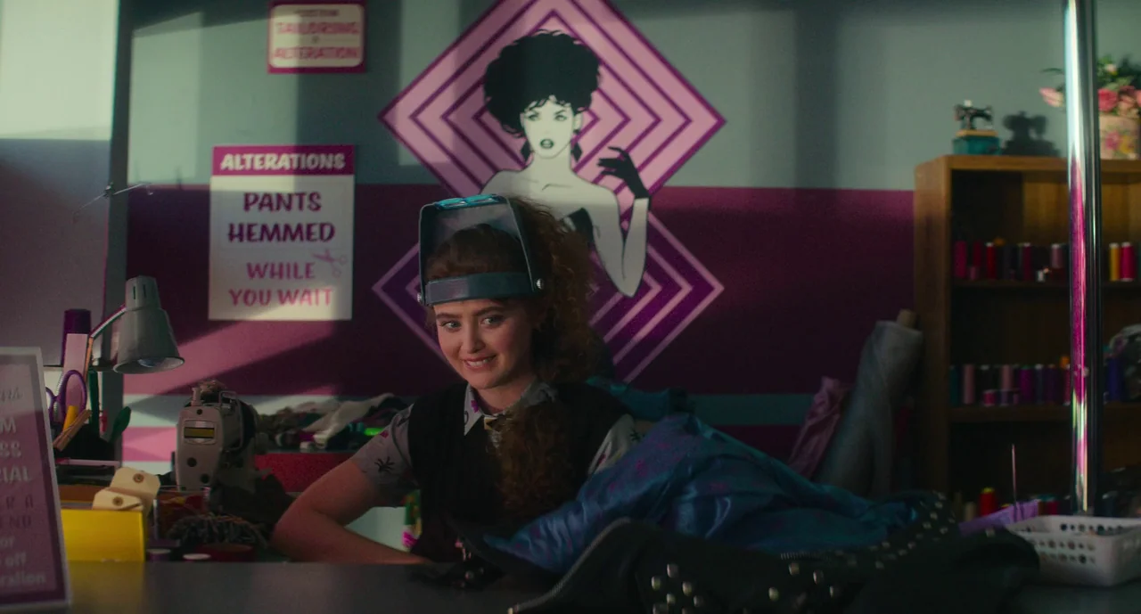 Lisa Frankenstein (2024) movie still: medium — A tailor shop workspace features wall signage and a stylized mural of a woman. A young woman wearing…