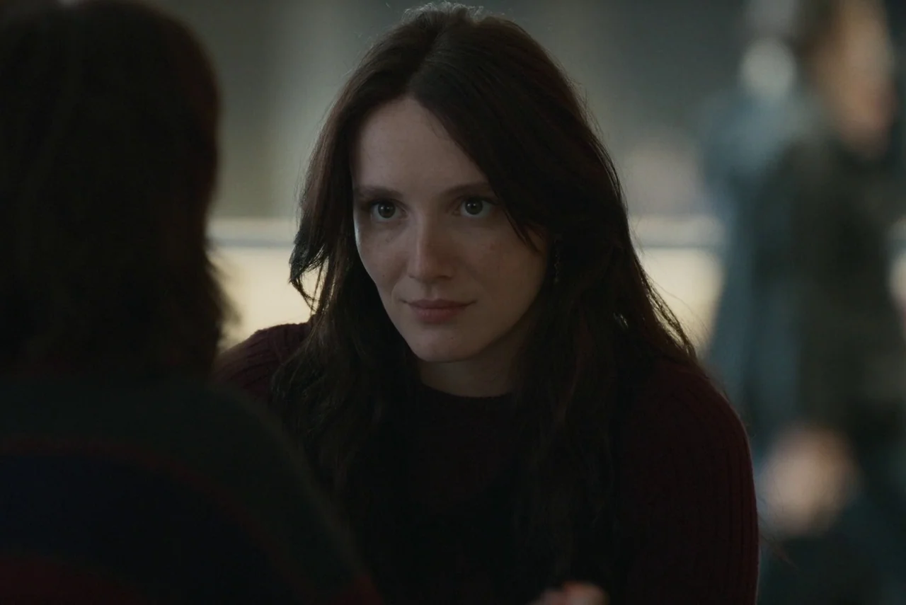 Red Rooms (2023) movie still: closeup — A dimly lit indoor space with blurred background figures visible behind the main subject. The woman in the…