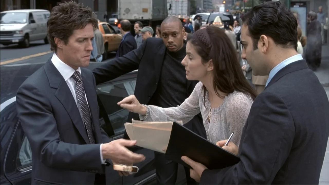 Two Weeks Notice (2002) movie still: medium — A busy urban street corner features a black car parked near a traffic lane. Three individuals stand…