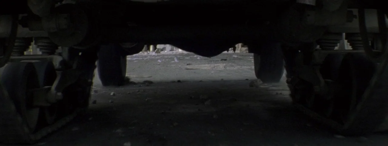 Post Mortem (2010) movie still: establishing — A view from beneath a heavy-duty transport vehicle with continuous track wheels. The ground is a dirt…