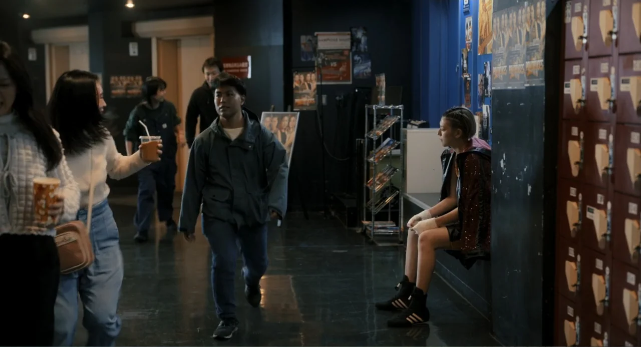 Knuckle Girl (2023) movie still: fullbody — A dimly lit indoor facility featuring rows of brown lockers and posters on walls. A man walks down the…