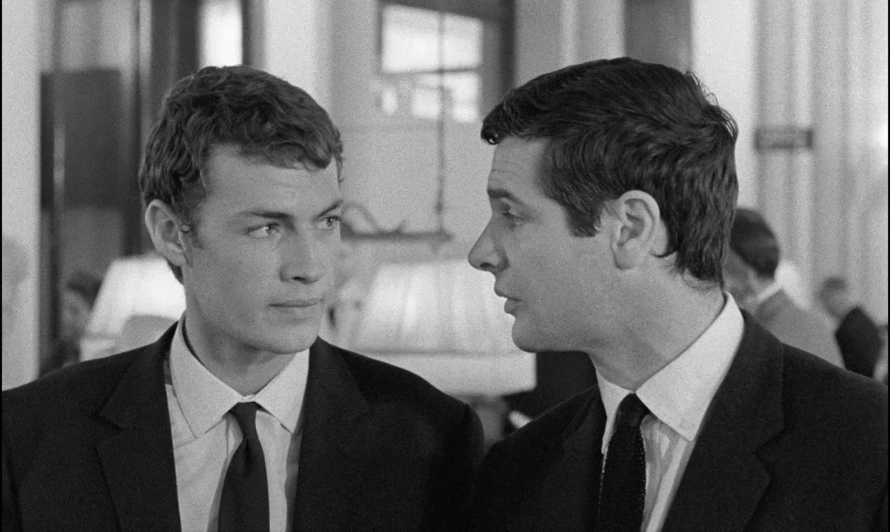 Bay of Angels (1963) movie still: medium — A bright interior space features table lamps in the blurry background. Two men in formal business attire…