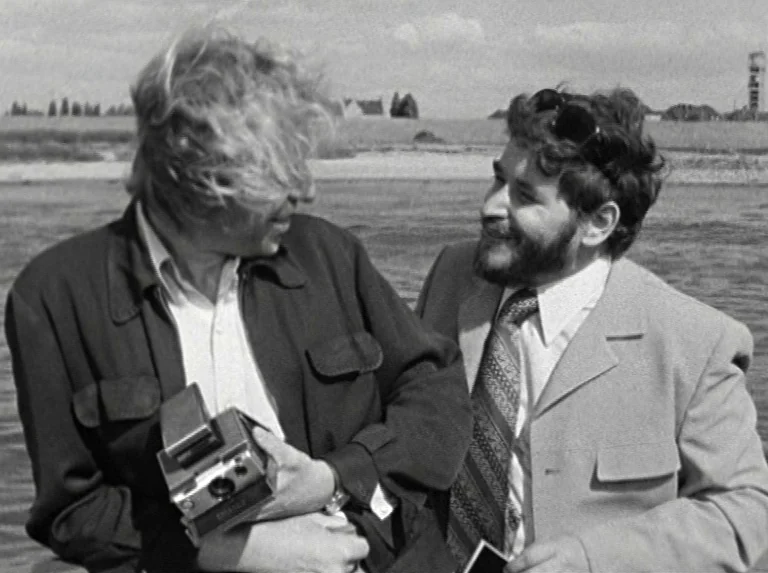 Alice in the Cities (1974) movie still: medium — Two men stand outdoors near a body of water under a bright sky. One man holds a vintage Polaroid…