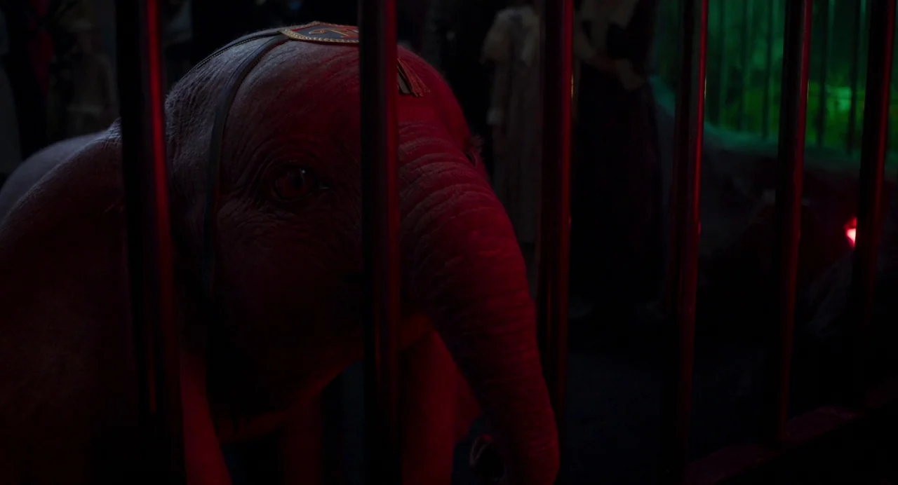 Dumbo (2019) movie still: closeup — A baby elephant is confined inside a dark, barred enclosure. The creature peers through the vertical bars under…