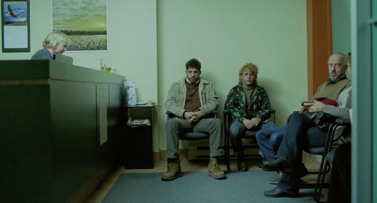 Tom at the Farm (2013) movie still: wide — A quiet office waiting area with pale green walls and a dark reception desk. Three men sit on black…