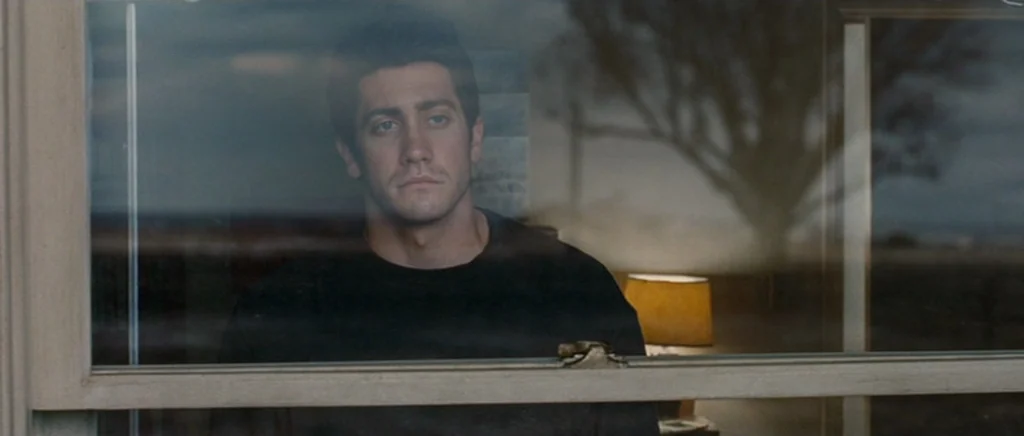 Jarhead movie still: medium — A man stands indoors looking out through a window pane during dusk. The reflection of a leafless tree is visible on…