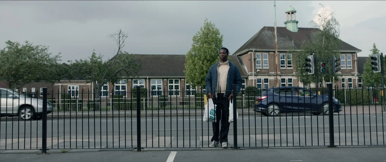His House (2020) movie still: fullbody — An asphalt road runs in front of a brick building with a green roof turret. A man stands holding two white…