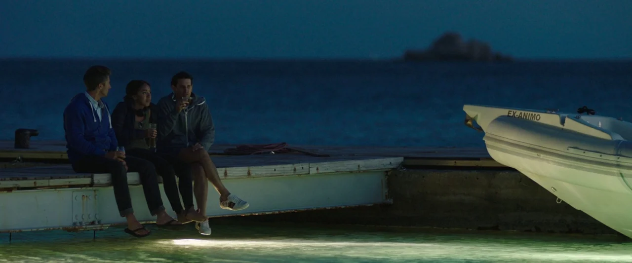 Greed (2019) movie still: wide — A wooden pier extends into the dark water at night under a blue sky. Three people sit along the edge holding drinks…