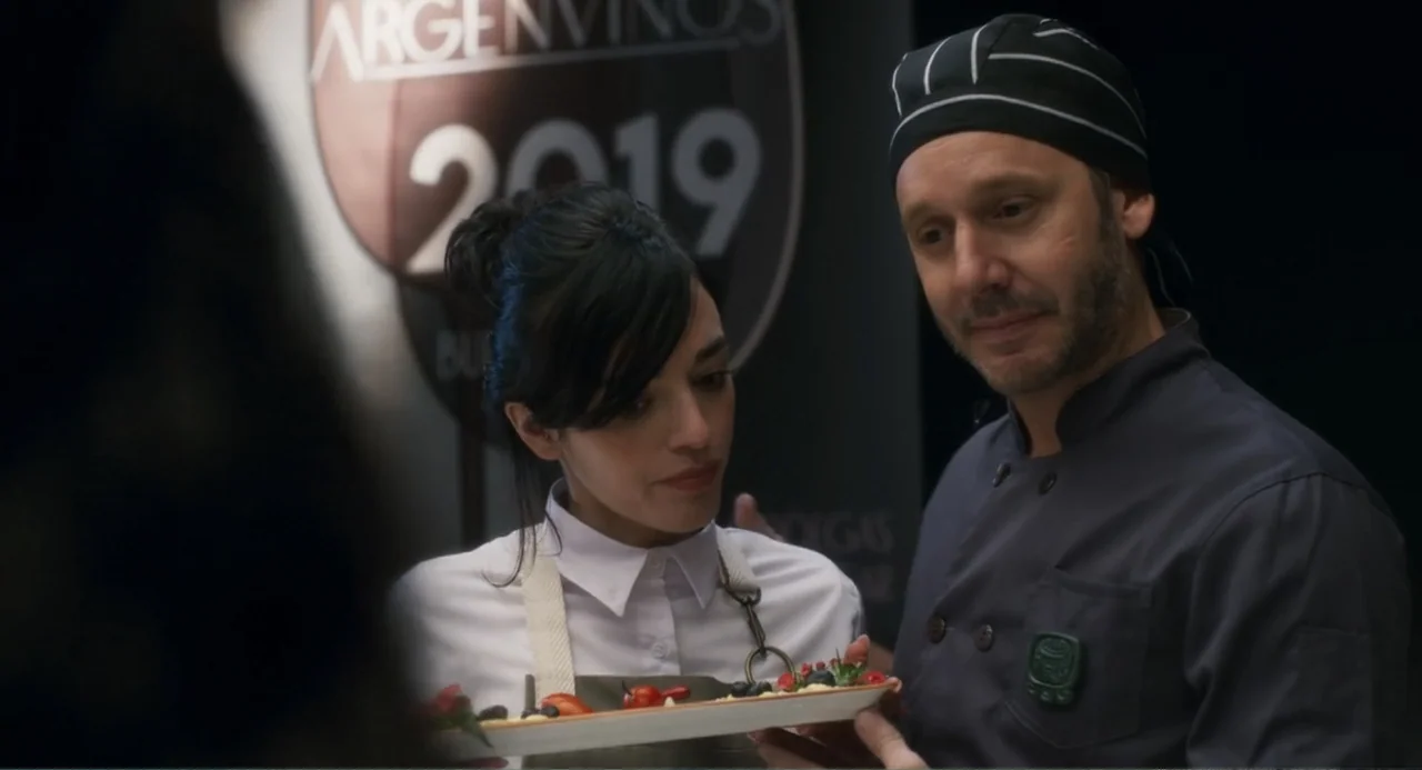 Message in a Bottle (1999) movie still: medium — A professional culinary display area with a branded banner in the background. A man and a woman in…