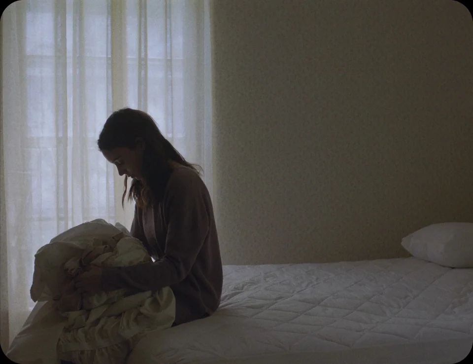 A Ghost Story (2017) movie still: medium — A minimalist room contains a bed with a white quilted cover and a curtained window. A woman with long…