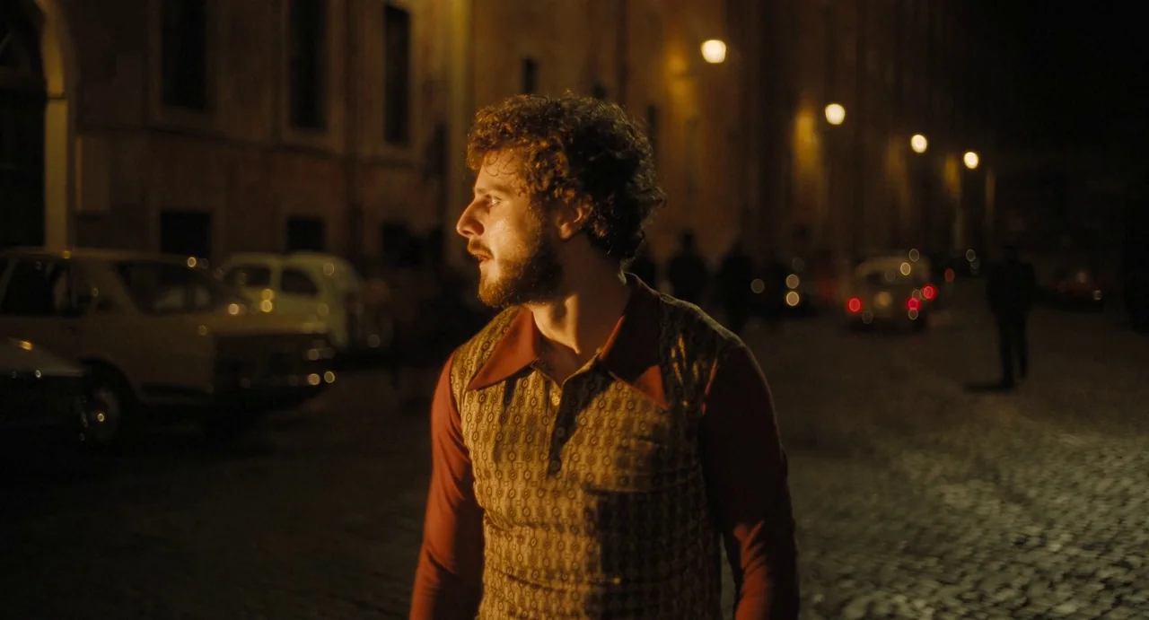 The First Omen (2024) movie still: medium — A quiet urban street at night with cobblestone paving and classic parked cars. A man with curly hair and…