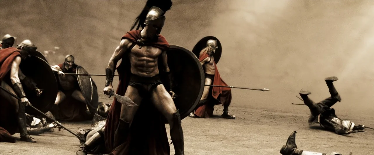 300 (2007) movie still: fullbody — A dusty, sun-drenched battlefield filled with Spartan soldiers in bronze helmets and crimson capes. Several…