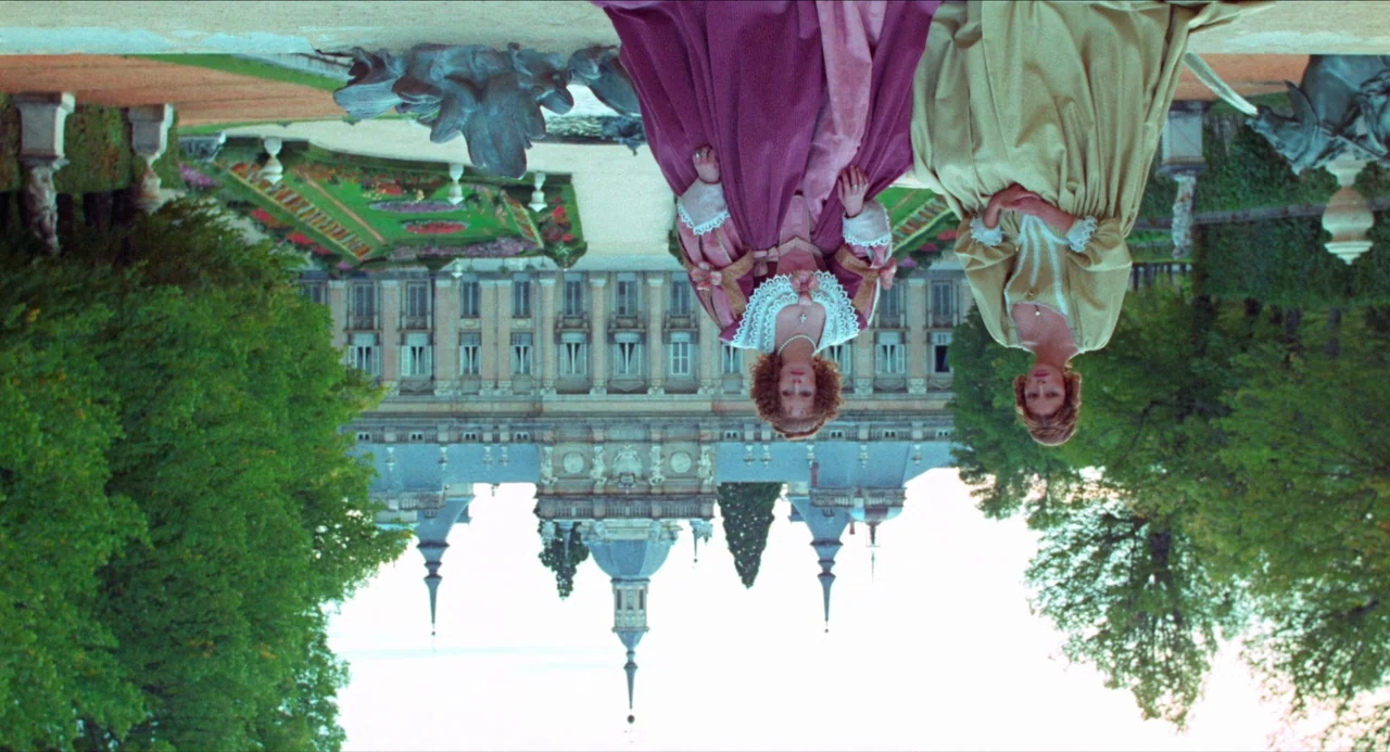 The Three Musketeers (1973) movie still: wide — Two women in historical gowns stand on the path of a manicured European estate. A large stone…