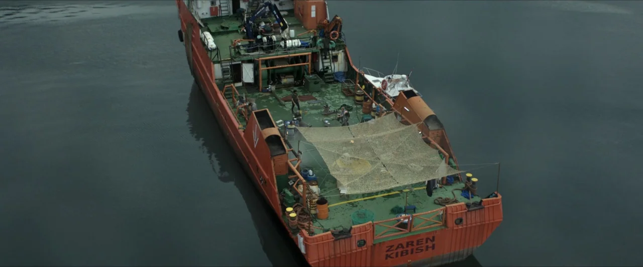 Apocalypse Z: The Beginning of the End (2024) movie still: wide — An orange industrial cargo vessel sits in calm dark water under an overcast sky.…