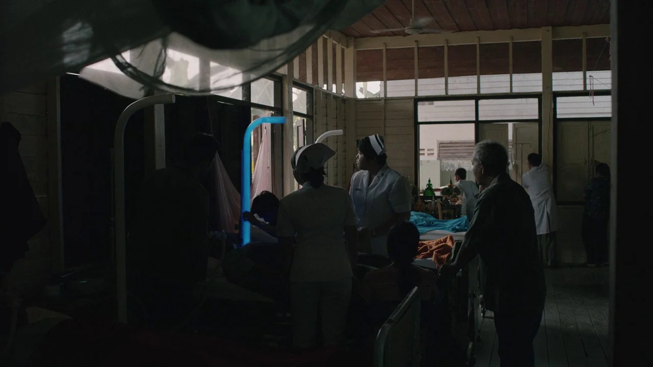 Cemetery of Splendour (2015) movie still: wide — A dimly lit room with wooden walls and large windows houses several hospital beds. Nurses and…