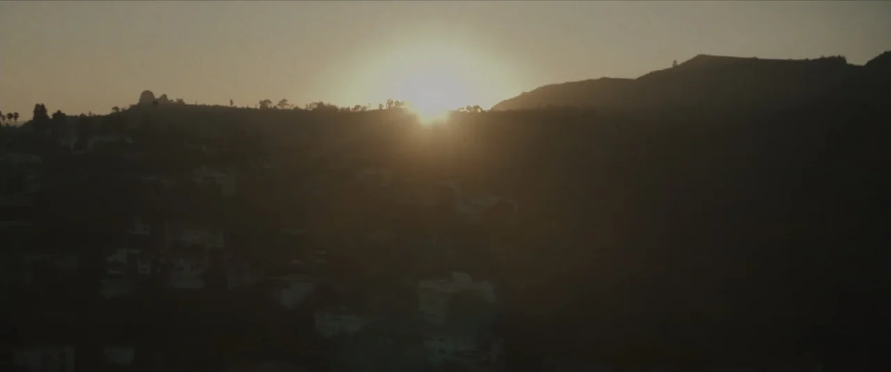 Stalker (2020) movie still: establishing — A view of rolling hills in the late afternoon with houses tucked away. The sun sets behind a mountain…