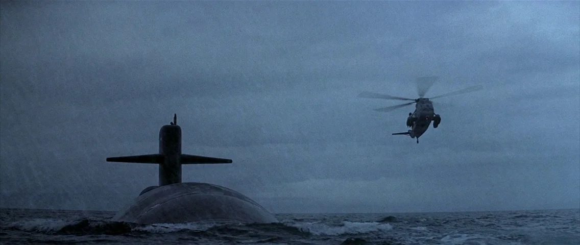 The Hunt for Red October (1990) movie still: wide — A dark military submarine partially emerges from the turbulent surface of the grey ocean. A…