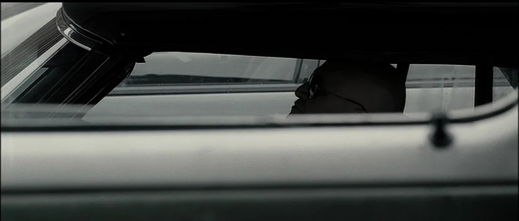 The Machinist (2004) movie still: closeup — The frame captures a view through the narrow window gap of a car door. A bald man wearing glasses sits…