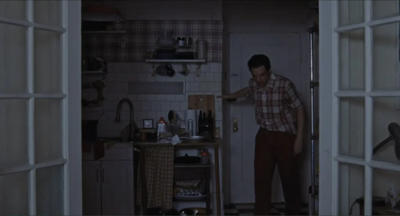 A Different Man (2024) movie still: medium — A dimly lit kitchen features plaid wallpaper and white tile backsplashes with cluttered countertops. A…
