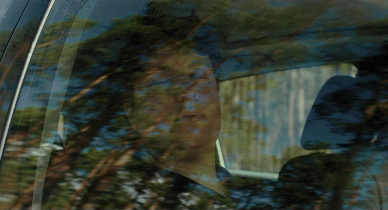 Afire (2023) movie still: closeup — The face of a young man is visible through a car window reflecting trees. He sits inside the vehicle while the…