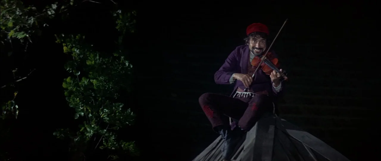 Fiddler On The Roof (1971) movie still: fullbody — A man sits on the peak of a dark pitched roof at night. Foliage is visible on the left side while…