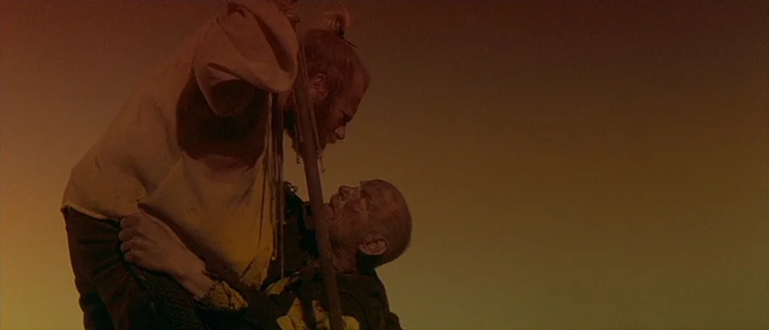 Flesh+Blood (1985) movie still: medium — An open field under a dusk sky with an expansive, gradient orange background. A man supports another man…