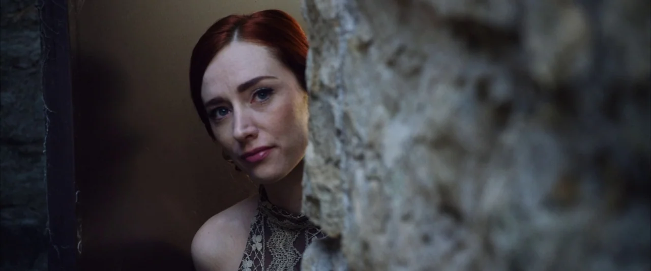 The Stylist (2020) movie still: closeup — A person looks out from behind a textured, weathered stone wall in an outdoor setting. The woman with red…