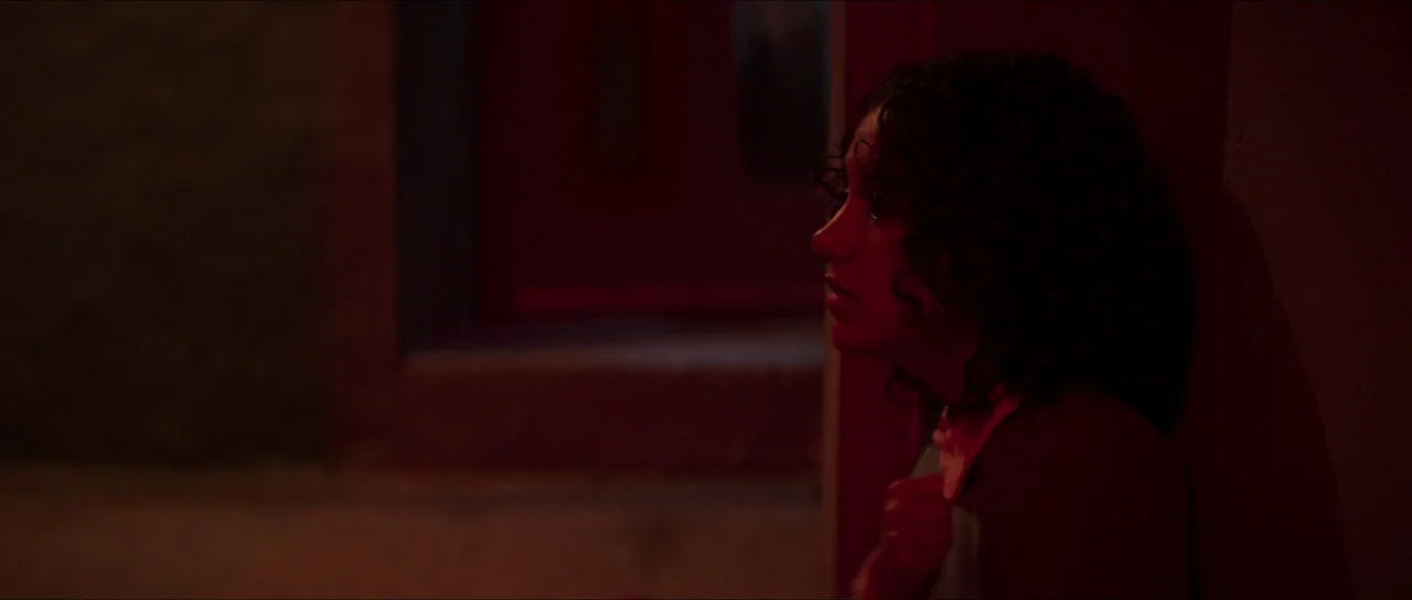 The Sacrifice Game (2023) movie still: closeup — A woman stands in the shadows of a dark outdoor space at night. She is illuminated by a harsh red…