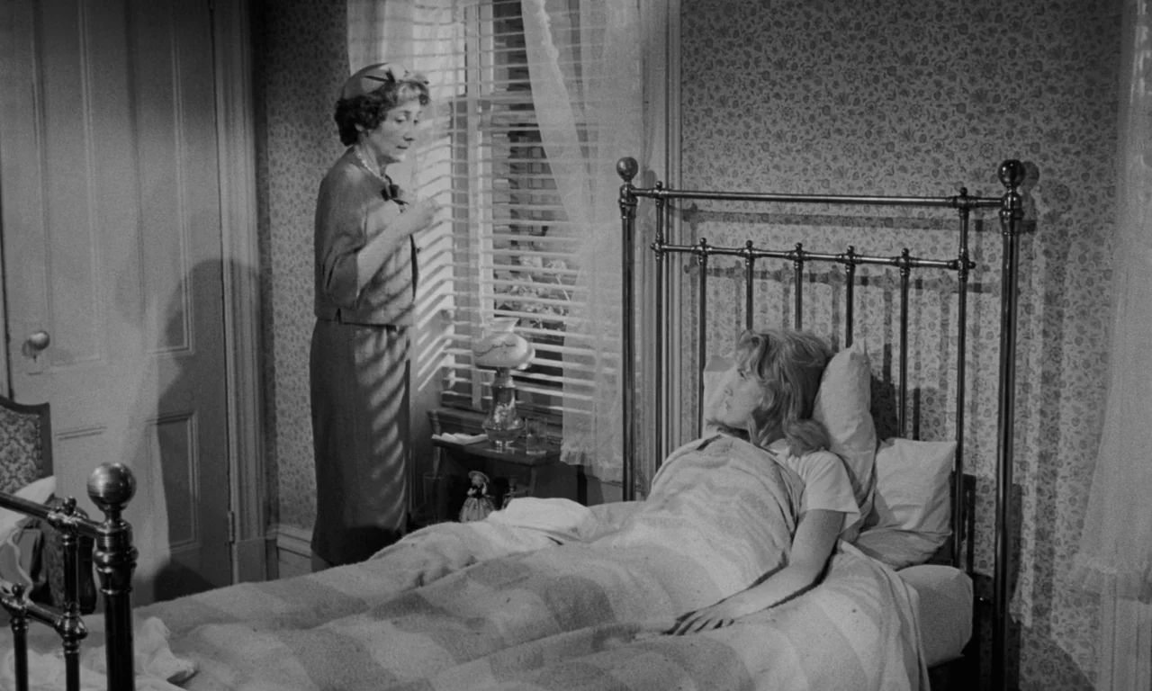 Something Wild (1961) movie still: medium — A traditional bedroom features floral wallpaper, a metal bed frame, and window blinds. An older woman…