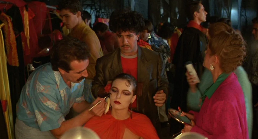 Matador (1986) movie still: medium — A crowded backstage dressing room with costumes hanging on racks in the background. A makeup artist works on a…