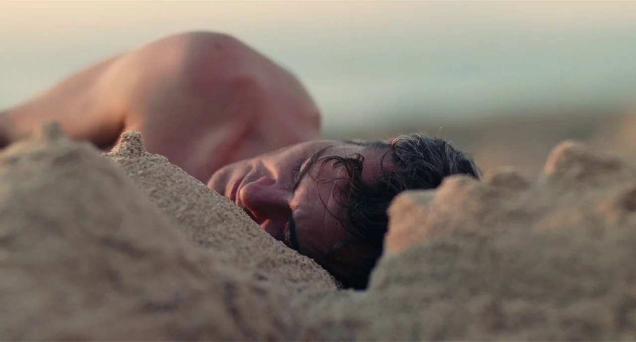 The Master (2012) movie still: closeup — A sandy beach at golden hour features a man partially buried in the sand. The man lies prone with his head…