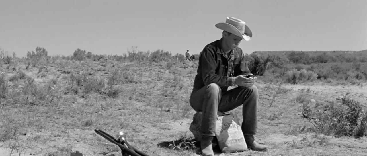 Hud (1963) movie still: fullbody — An arid desert plain stretches toward a distant ridge under a bright sky. A young man sits on a stone with a bag,…