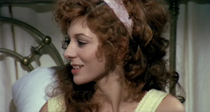 Blood For Dracula (1974) movie still: closeup — A woman rests against a metal bed frame covered by white linens. She sits upright in bed while…