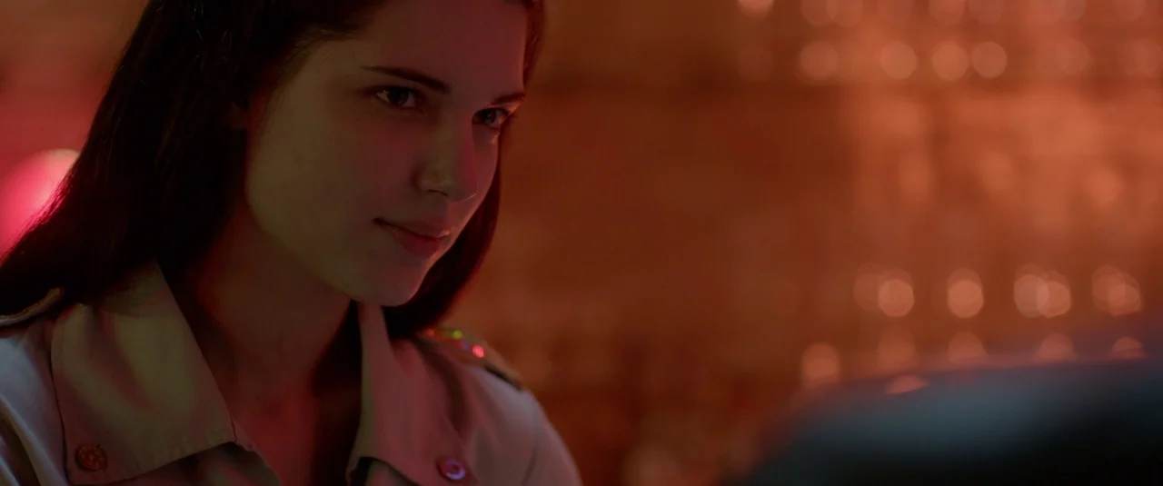 The Lure (2015) movie still: closeup — A dark indoor space with blurred, out-of-focus bokeh lights visible in the background. A woman in a tan shirt…
