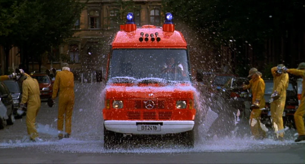 Run Lola Run (1998) movie still: wide — A bright red emergency van drives forward through a thick spray of white foam. Several people in yellow work…