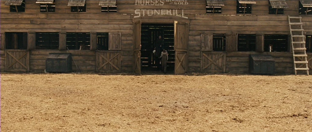 True Grit (2010) movie still: wide — A rustic wooden stable building features horizontal log planks and multiple stalls with doors. A long ladder…