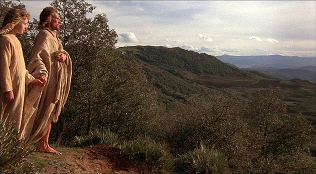 The Last Temptation Of Christ (1988) movie still: fullbody — Two individuals stand on the edge of a grassy, tree-lined ridge overlooking a vast…