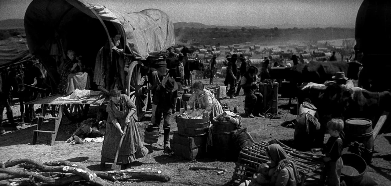 The Big Trail (1930) movie still: wide — A vast, dusty prairie encampment is filled with covered wagons and groups of people. Settlers in period…