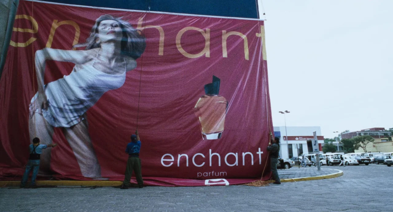 Amores Perros (2000) movie still: wide — A sprawling outdoor parking lot contains a massive red vinyl perfume advertisement banner. Three workers…