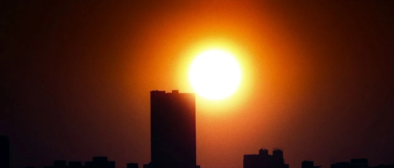 Archenemy (2020) movie still: establishing — A city skyline is silhouetted against a brilliant orange and red sunset sky. A tall rectangular…