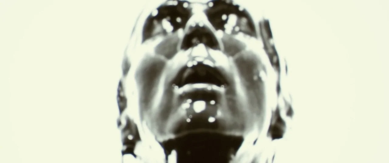 Beyond The Black Rainbow (2010) movie still: closeup — A metallic face is illuminated against a blindingly bright white background. The subject is…