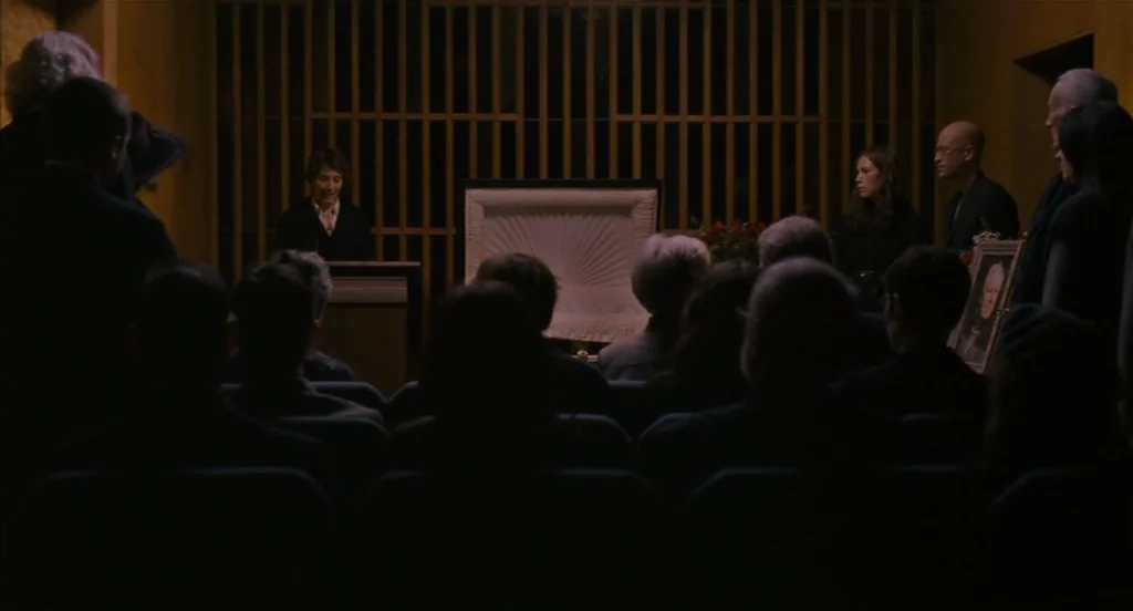 Restless (2011) movie still: wide — A dimly lit room features a wooden-slatted wall behind an open white casket. A woman addresses a group of…