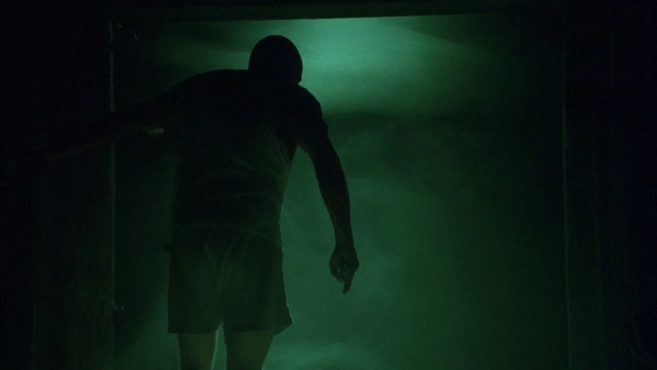Saw movie still: fullbody — A dark, shadowed space is defined by a doorway filled with intense green light. A man stands in the foreground, his back…
