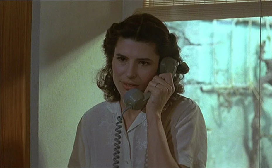 The Woman Next Door (1981) movie still: medium — A woman stands indoors near a window with light filtering through a blind. She is speaking on a…